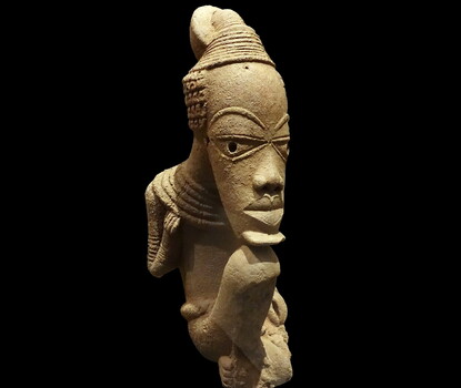 A stylized terracotta sculpture of a seated man with an oversized and elongated head relative to their body, chin resting on the figure's knees. Damage to the figure's shins.