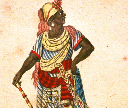 A Black man in a multicolored robe and cape, a gold hat with a red frill, and silver earrings, looking upward.