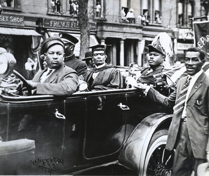 An open-topped car with four passengers in a parade. The driver is a Black man with a dark jacket, white shirt, dark tie, and dark driver's cap. In the back seat, from left to right, is a Black man in a military uniform; a Black man wearing academic regalia; Marcus Garvey, a Black man, wearing a military uniform and holding a sword. Next to the car is a Black man wearing a dark jacket, white shirt, and striped tie. In the background are apartment buildings, where Black people are looking on at the parade.