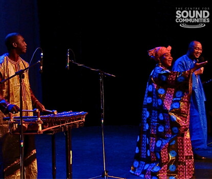 From left to right: Fodé Lassana Diabaté, a Black man wearing a light-colored tunic and dark red shirt, playing the balafoon (a struck instrument similar to a xylophone); Hawa Kassé Mady Diabaté, a Black woman wearing a patterened cloth dress and red headscarf, singing with arms outstretched, and Chérif Keita, a Black man in a blue robe, smiling.