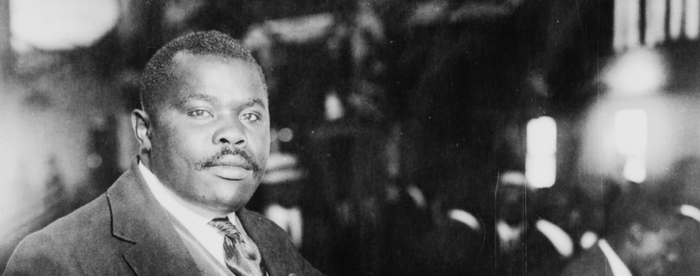 A black-and-white photograph of Marcus Garvey, a Black man wearing a dark suit, light-colored vest, white shirt, and dark tie, sitting in a wooden chair next to a desk filled with papers, holding a handkerchief in his right hand and a gavel in his left.